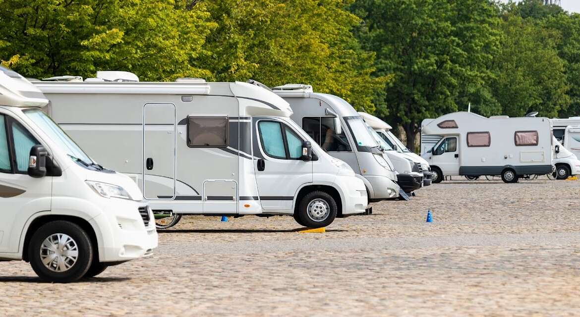 Row of white motorhomes parked on a cobblestone lot next to a line of green trees.