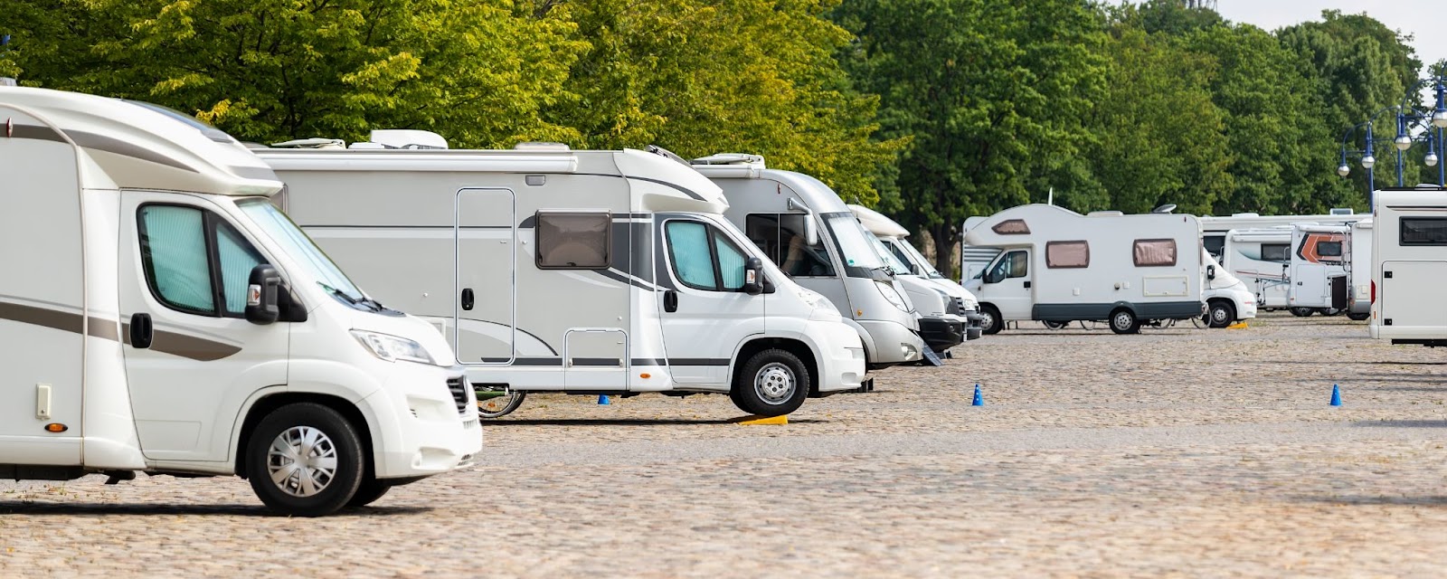 Row of white motorhomes parked on a cobblestone lot next to a line of green trees.