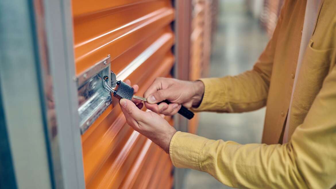 A person in a yellow jacket locks an orange storage unit with a padlock using a key. The corridor has a row of similar storage units, suggesting a secure storage facility.