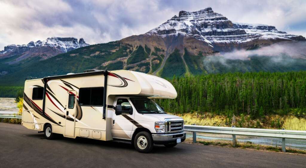 White RV parked on a road with snow-capped mountains and forest in the background.