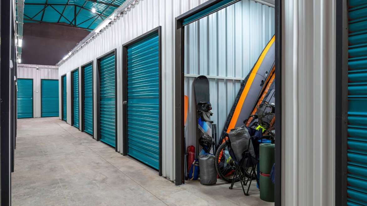A row of teal storage units with one open, revealing sports equipment like a snowboard, paddleboard, and camping gear inside.