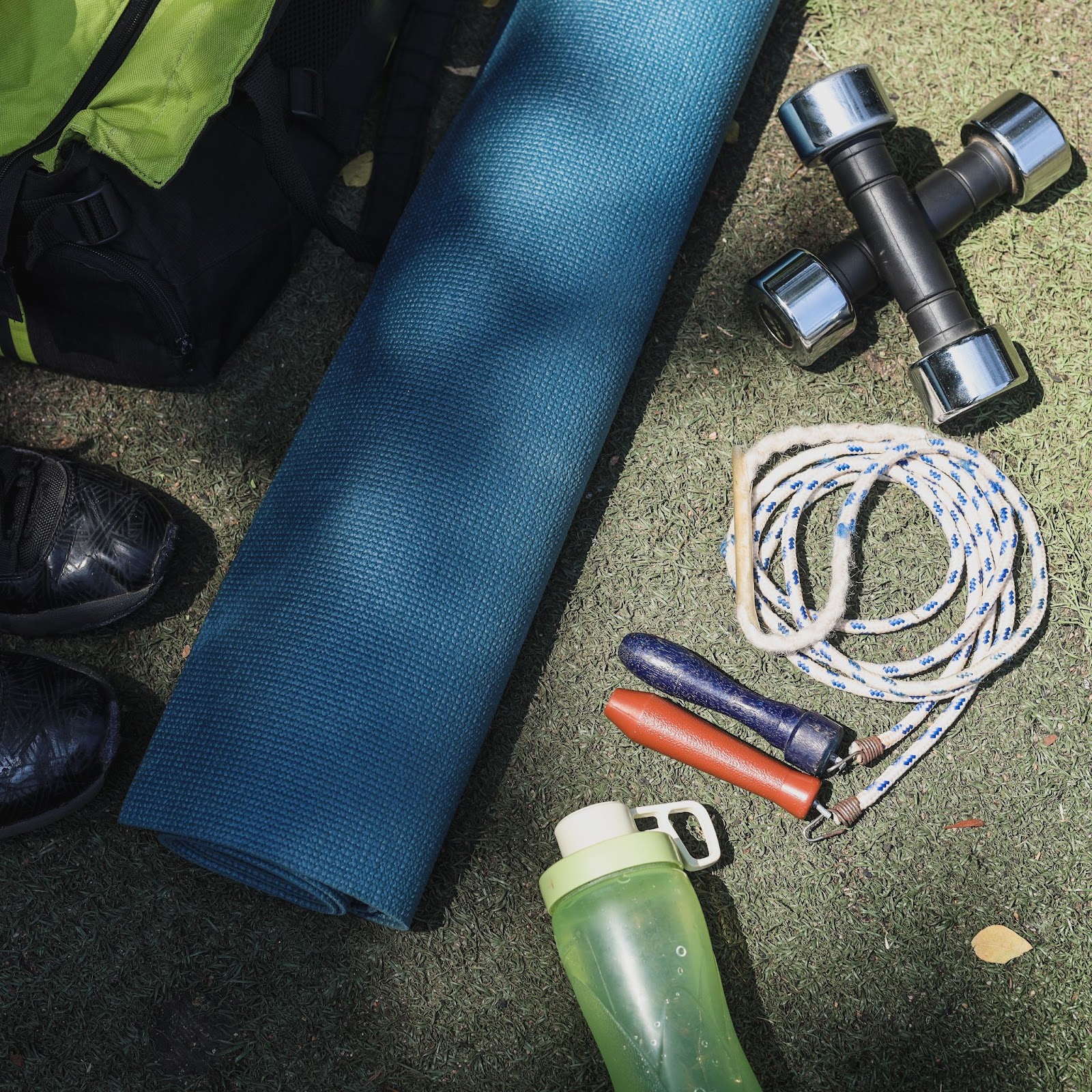 Fitness gear laid out on grass for an outdoor workout rolled blue yoga mat, pair of small chrome dumbbells, jump rope, water bottle, running shoes, and gym bag.
