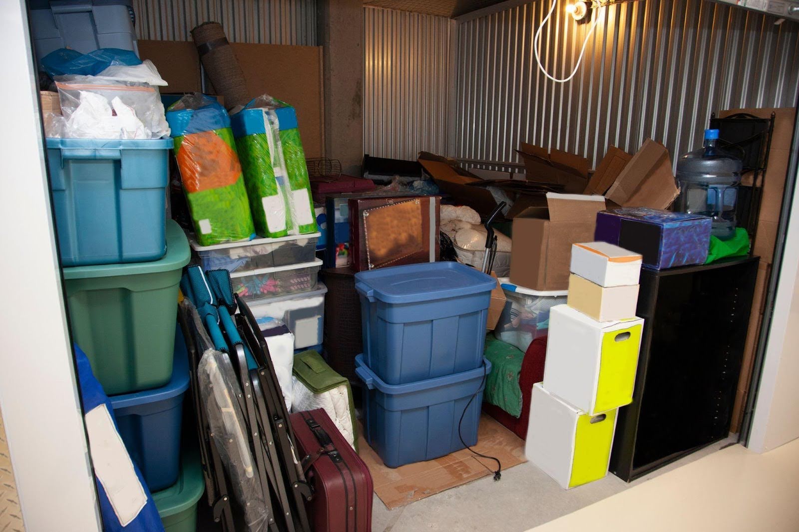 A cluttered storage unit filled with blue plastic bins colorful boxes folded furniture and a water dispenser