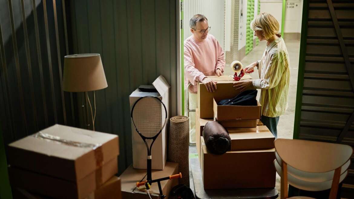 A couple packing boxes in a storage unit with items like a lamp and tennis racket