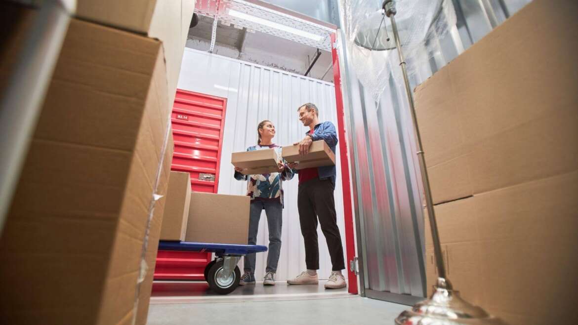 A man and woman are lifting boxes together inside a storage unit organizing their belongings