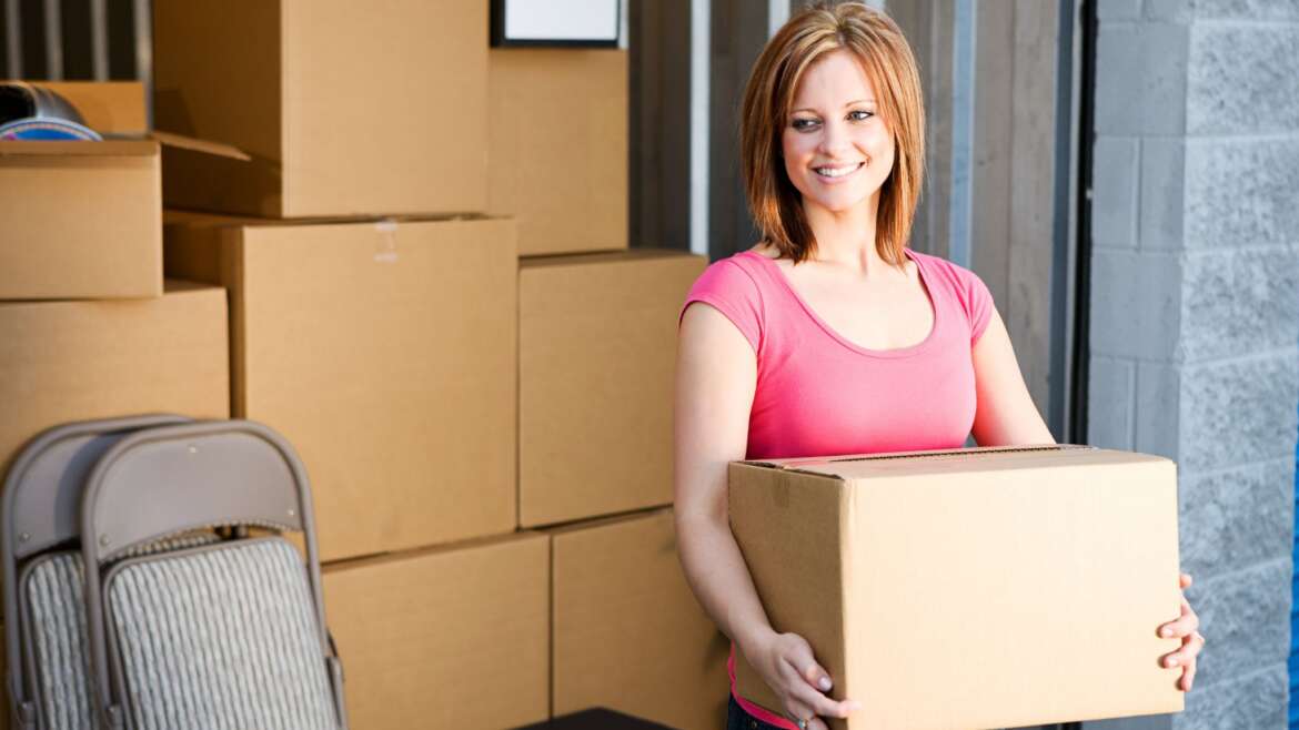 A smiling woman carrying a moving box in front of a stack of cardboard boxes.