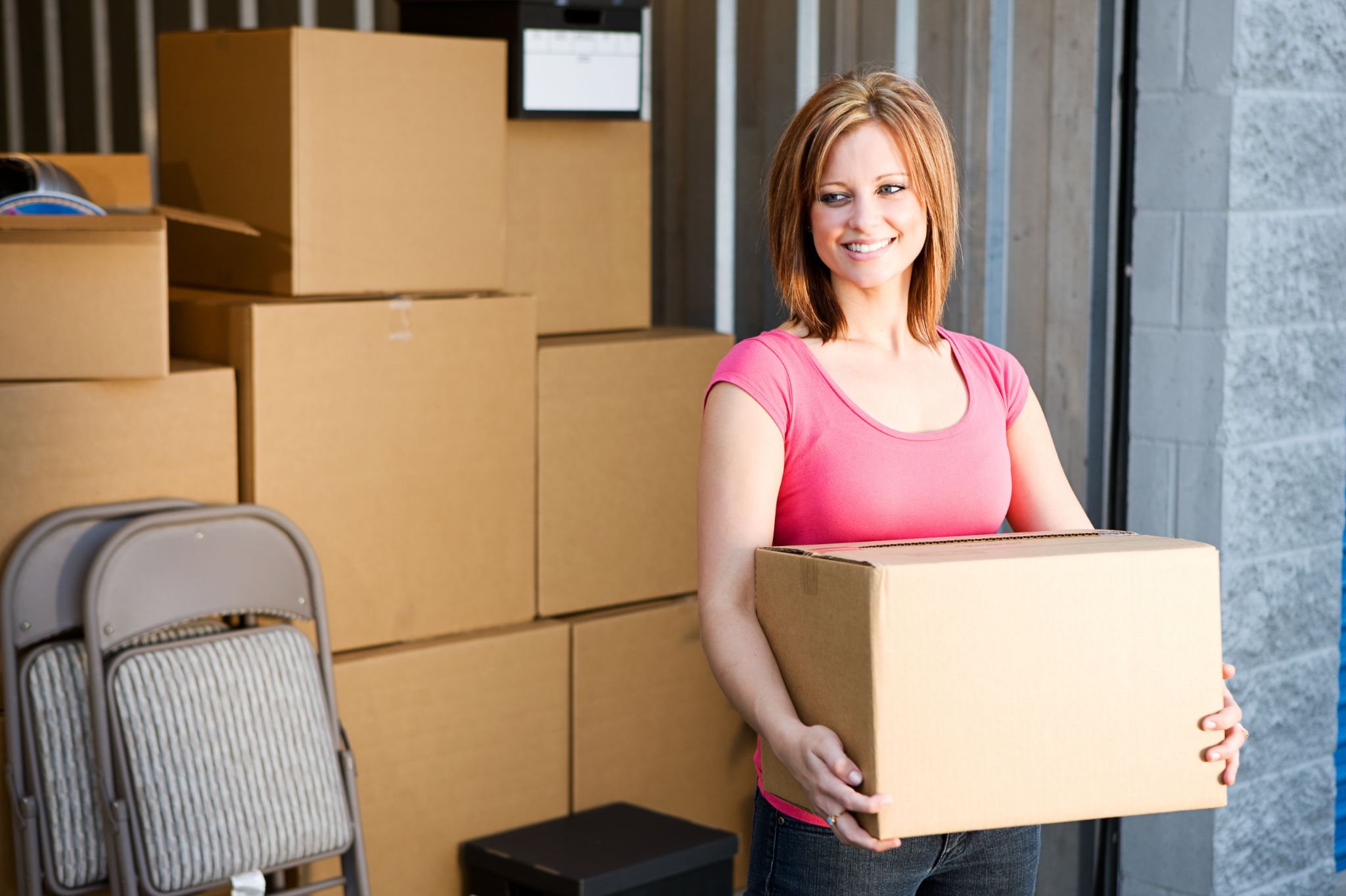 A smiling woman carrying a moving box in front of a stack of cardboard boxes.