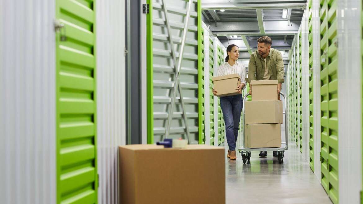 A man and woman navigate a storage unit filled with stacked boxes and miscellaneous items